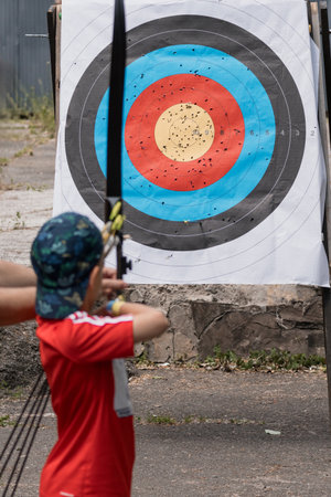A young archer in a red shirt practices shooting target with a bow and arrow outside, concentrating...