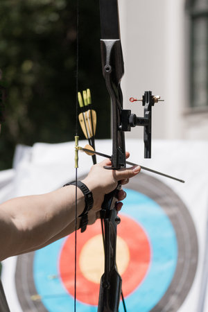 Focused young archer practices archery outdoors with a black recurve bow, aiming at a colorful...