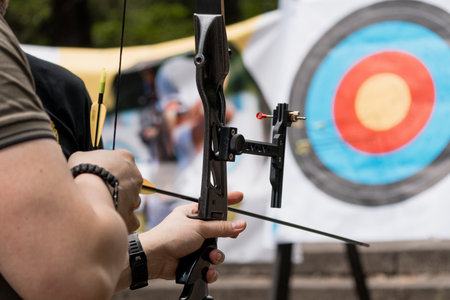 Focused male archer intensely draws a compound bow, aiming for a blue-ringed target during an outdoor archery practice session, showcasing sport and precisionの写真素材