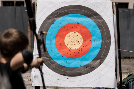 Focused boy practices archery outdoors, aiming at a target board densely covered with blue and red arrow hits, utilizing a wooden stand. recreational sport sceneの写真素材