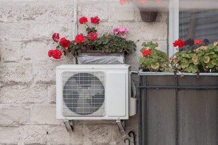 A white air conditioner is mounted on a light gray stone wall below a window box overflowing with vibrant red geraniums, showcasing home cooling with garden decorの写真素材