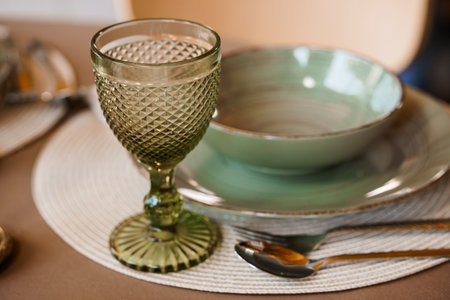 A beautifully arranged table setting features a green glass goblet and matching ceramic bowl on a woven placemat, golden cutlery, and warm brown tones. evokes rustic eleganceの写真素材