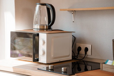 A clean kitchen corner features a white microwave stacked atop a wooden surface, alongside a glass electric kettle and wall outlets, evoking a bright, modern lifestyle aestheticの写真素材
