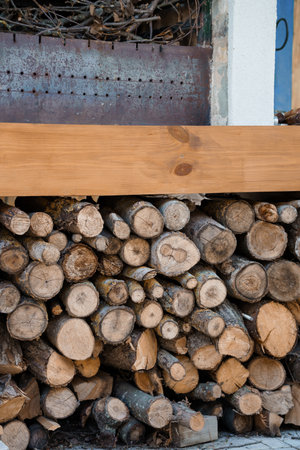 A neatly stacked woodpile against a weathered metal panel features a light wooden beam resting atop,...