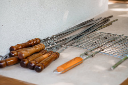A close-up shot displays an assortment of stainless steel meat skewers with warm wooden handles and a silver grill grate resting on a light-colored countertop for outdoor cookingの写真素材