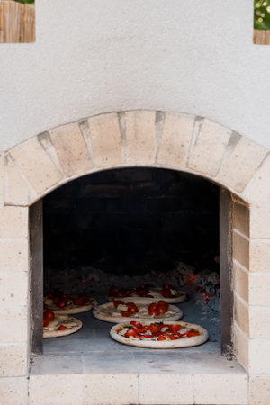 A traditional stone pizza oven bakes several margherita pizzas inside, displaying the art of Italian cooking in an outdoor kitchen setting with warm lightingの写真素材
