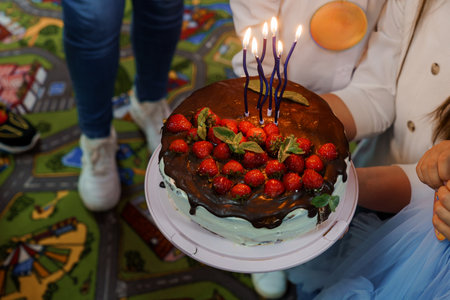 A close-up shot of a delicious chocolate strawberry cake with lit candles held by two people during a cheerful birthday party, capturing a festive mood and joyful celebrationの写真素材