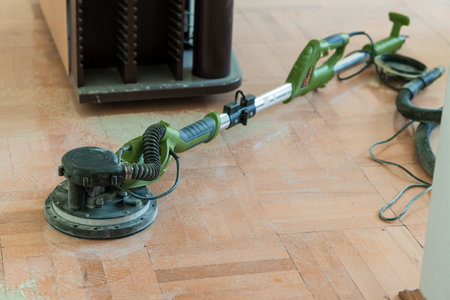 A green power sander is being used to refinish a wooden floor in a brightly lit interior room, with a dust vacuum hose connected for immediate dust extractionの写真素材