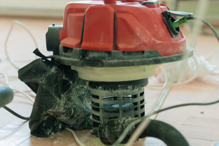 A close-up shot of a red workshop vacuum cleaner covered in dust, situated on a light wooden floor...
