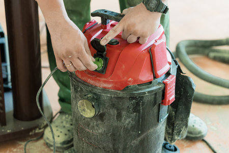 A person in olive green clothing connects a green plug into a red shop vacuum cleaners port, illustrating industrial cleaning in a workshop settingの写真素材