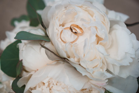 Close-up shot of gleaming gold wedding bands resting on a pristine white peony bouquet, representing romance, fidelity, and a joyful marriage ceremonyの写真素材