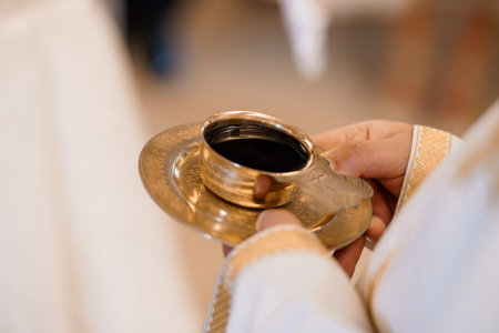 A close-up image showing a priest holding a golden chalice filled with red wine during a Catholic Eucharist ceremony. depicts faith, devotion, worship, and religious traditionの写真素材