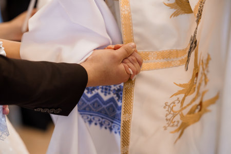 A close-up shot of two hands clasped in prayer during a wedding ceremony, with the groom's hand wearing a dark suit and the priest's in an embroidered vestmentの写真素材