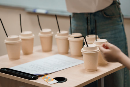 A close-up depicts shots an array of white paper cups set on a light wooden table for beverage sampling, with a hand reaching for one, promoting product testingの写真素材