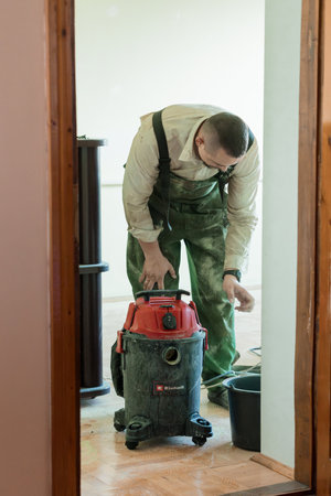 A young man, covered in dust, cleans a hardwood floor with a red Einhell vacuum cleaner during a home renovation project inside a houseの写真素材