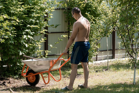 A shirtless man pushes an orange wheelbarrow through a lush garden yard on a sunny summer day,...