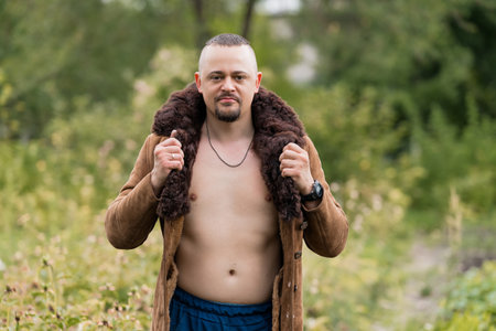 An intense outdoor portrait showcases a muscular, bearded man, with an open brown suede coat...