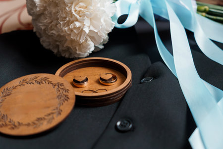 Close-up shot of wedding bands presented on an engraved wooden ring box resting on a black suit, adorned with white flowers and a blue ribbonの写真素材