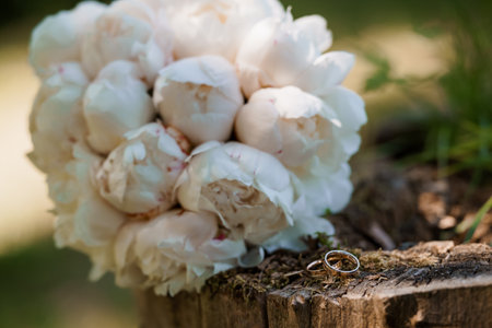 A close-up shot showcases elegant wedding bands resting atop a rustic wooden log alongside a lush bouquet of white peonies, symbolizing love and commitmentの写真素材