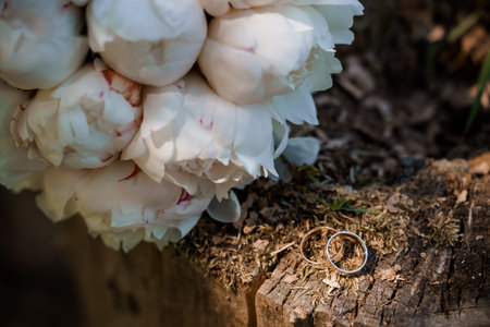 A lush white peony bouquet rests near a pair of golden wedding rings placed on weathered tree bark, capturing a serene and intimate moment of loveの写真素材