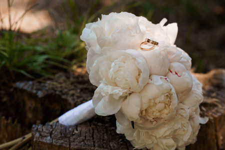 A beautiful close-up showcases elegant gold wedding rings resting gently on a lush white peony bouquet, evoking romance. perfect for wedding invitations, engagement announcements, or bridal themesの写真素材