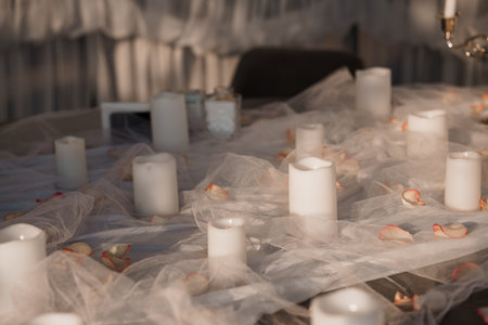 A softly lit indoor setting features a table draped with white tulle and scattered rose petals, adorned with multiple candles creating a romantic ambiance for couplesの写真素材