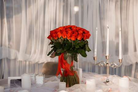 A large bouquet of vibrant red roses is beautifully arranged in a glass vase with a red ribbon, alongside elegant white candles on a draped table setting, symbolizing romance and celebrationの写真素材