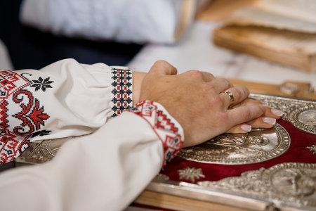 A close-up shows hands clasped in prayer over a richly decorated Orthodox Christian holy book, featuring a golden cover and red textile, while the sleeves display intricate Ukrainian embroideryの写真素材