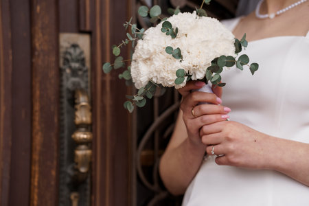 A beautiful bride with a wedding band holds a lush white hydrangea bouquet complemented by...