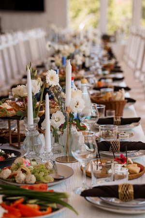 A long, elegantly decorated wedding table features a lavish spread of food, white floral arrangements, and detailed place settings, conveying a chic and celebratory moodの写真素材