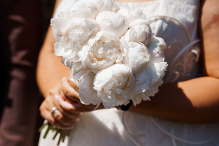 A close-up captures a brides hands delicately holding a lush bouquet of pure white peonies, symbolizing love and new beginnings, bathed in warm sunlight, romantic themeの写真素材