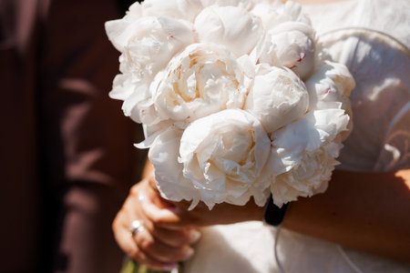 A close-up shot showcases a bride holding a stunning bouquet of white peonies, suggesting an elegant wedding theme with soft lighting and a focus on floral beautyの写真素材