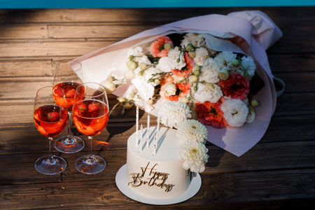 A beautiful birthday still life features a white cake with Happy Birthday lettering alongside a lush bouquet, wine glasses filled with a pink drink, and candles, set on a rustic wooden tableの写真素材
