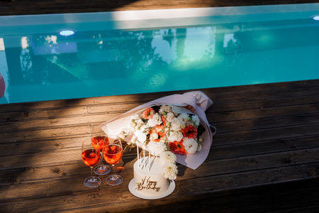 A romantic birthday setup with a white cake, roses, champagne glasses, and sparkling wine on a warm wooden deck overlooking a vibrant blue swimming poolの写真素材