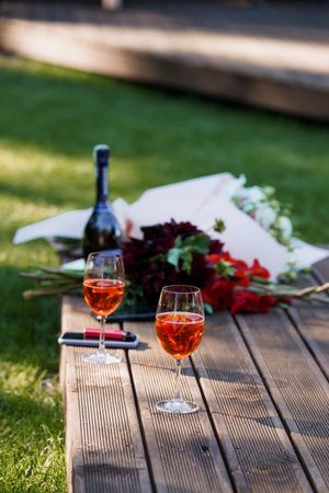 Intimate garden picnic featuring two glasses of rose wine with a bouquet of red roses on a rustic wooden table, evoking a summer evening moodの写真素材