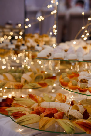 A beautiful tiered dessert arrangement overflows with colorful sliced fruits like apples, bananas, and oranges alongside delicate pastries, creating a luxurious ambiance illuminated by twinkling fairy lights, perfect for weddings, parties, or festive gatheringsの写真素材
