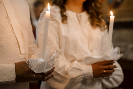 A close-up shot captures hands holding lit candles within an Orthodox Christian church setting, with individuals clad in white robes and golden rings, evoking a solemn religious moodの写真素材