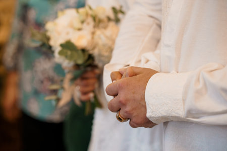 Wedding Ceremony Hands Clenched Together with Bouquet. White Flowers, Grooms Attire, Formal Event, Love, Joy, Blurred Background, Warm Tone.の写真素材
