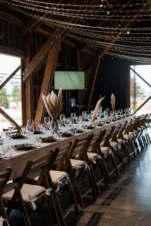 A long banquet table is elegantly set for a wedding reception inside a spacious rustic barn,...