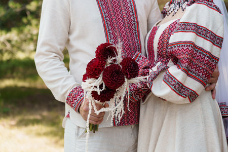 A beautiful Ukrainian couple in cream-colored embroidered wedding attire embraces lovingly, holding a vibrant bouquet of red dahlias outdoors, capturing a rustic, folk-style celebrationの写真素材