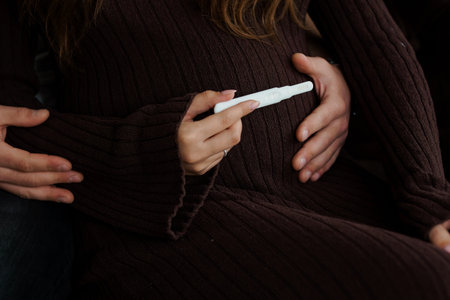 A loving couple gently holds a positive pregnancy test over a woman's baby bump, sharing a tender moment of joy and anticipation. brown knitwear and soft lighting create a warm, intimate moodの写真素材
