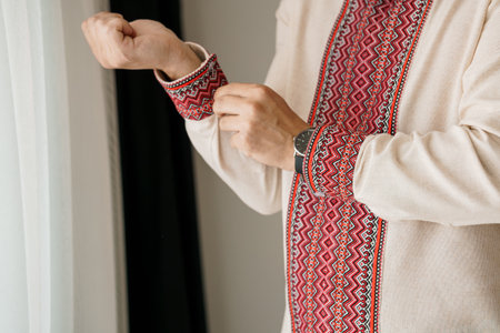 A close-up shot of a man carefully fastening the cuffs of a cream-colored Vyshyvanka shirt adorned...
