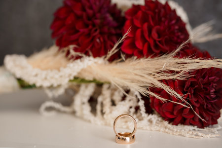 A close-up shot showcases elegant gold wedding rings resting against a lush bouquet of deep red dahlias accented with delicate pampas grass. symbolizing love, commitment, and a celebratory occasionの写真素材