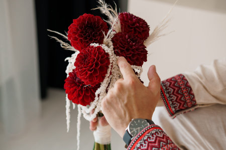 A close-up shot of a hand holding a vibrant red dahlia boutonniere adorned with delicate white feathers, paired with a traditionally embroidered sleeve, suggesting a Ukrainian wedding or cultural eventの写真素材