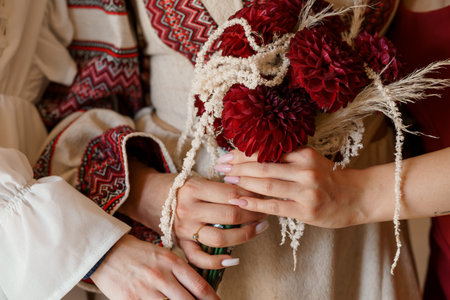 Two pairs of hands gently clasp a vibrant burgundy dahlia wedding bouquet, adorned with natural accents, showing Ukrainian embroidered shirts. intimate moment, cultural heritage, floral designの写真素材