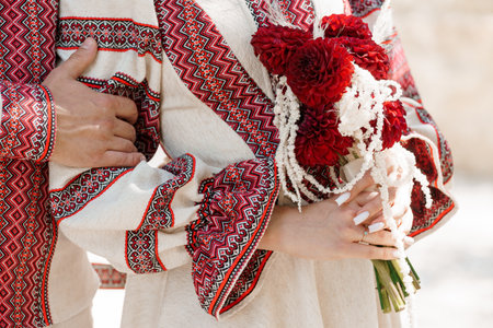 A close-up of a Ukrainian couple in intricately embroidered traditional wedding clothing holding a vibrant bouquet of deep red dahlias, symbolizing love and heritage, cultural eventの写真素材