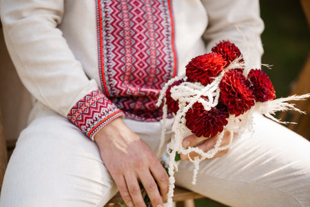 A close-up shot of a groom in traditional Ukrainian attire a cream vyshyvanka shirt with intricate red embroidery clutching a bouquet of deep red dahlias during a wedding ceremonyの写真素材