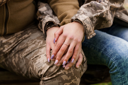 A close-up image showcasing hands clasped together, one wearing a military camouflage jacket and the other denim jeans, with a diamond ring visible, suggesting love, commitment, and partnershipの写真素材
