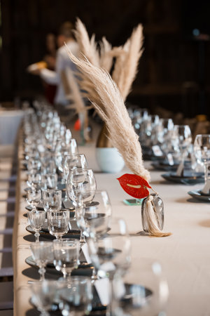 A long wedding table is beautifully set with glassware, plates, and unique floral centerpieces featuring pampas grass and vibrant red anthurium blooms. rustic, chic reception ambianceの写真素材
