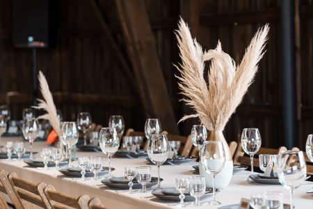 A beautifully arranged wedding reception table features delicate glassware, dark plates, and striking pampas grass in vases, set within a charming rustic barn interior for a sophisticated eventの写真素材